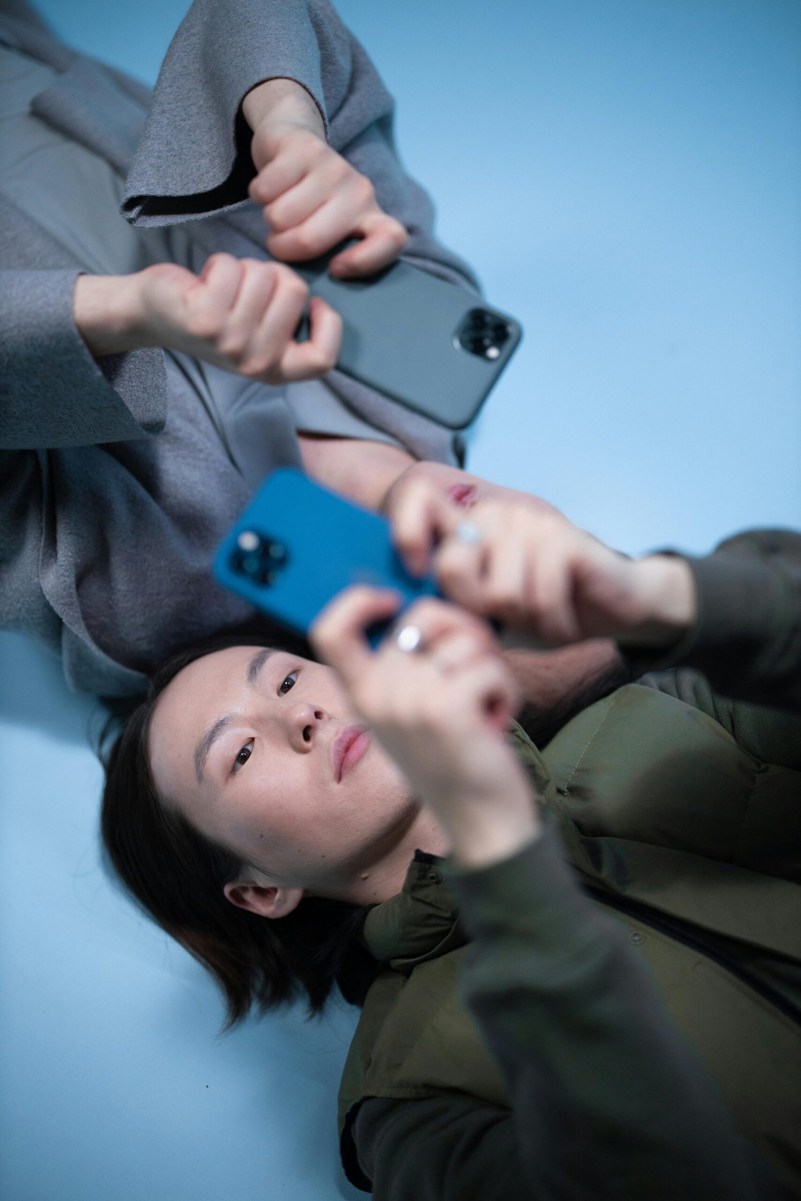 Two people lying down, using smartphones against a blue background indoors.